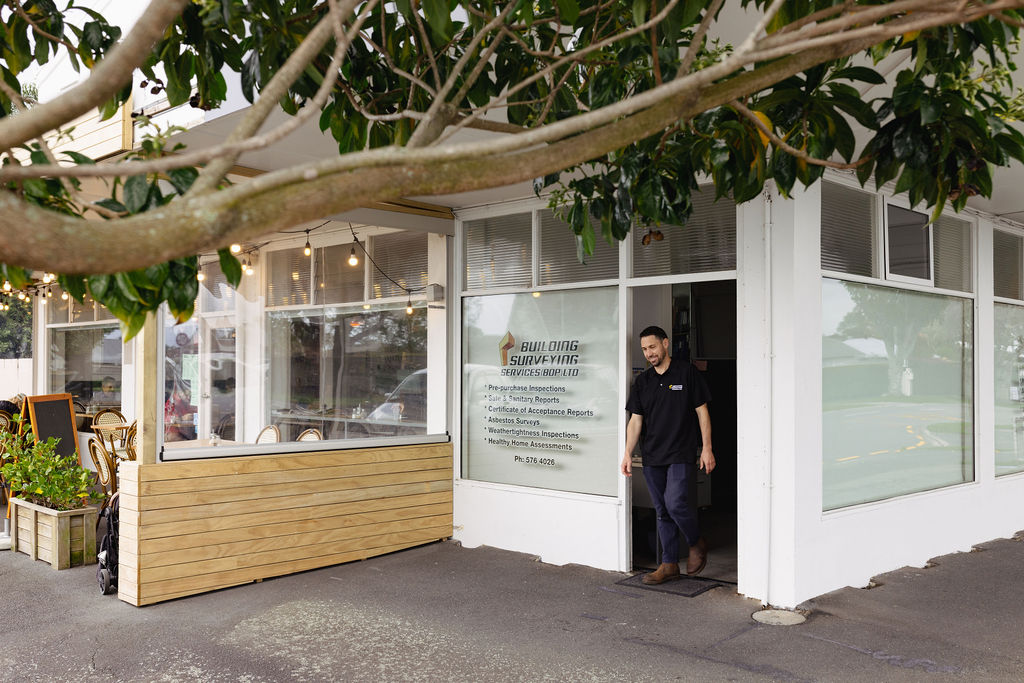 A man exits a building with large windows displaying "Building Surveying Services." Nearby is a café with string lights and outdoor seating.