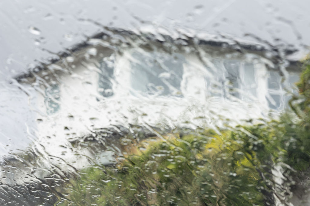 A blurred view of a white house with large windows seen through a rain-soaked window. Raindrops streak down, creating a distorted, moody effect.