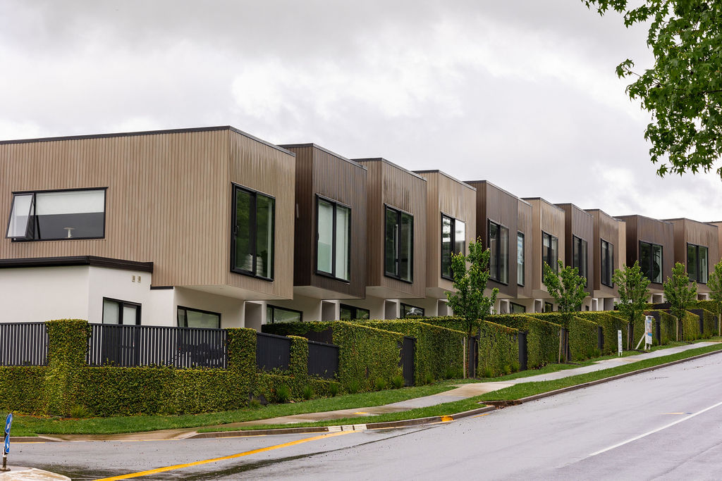 Modern row of townhouses with flat roofs and large windows, lined by hedges and trees on a sloped street, under a cloudy sky.